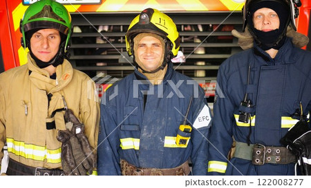Portrait of young happy fireguards in protective uniforms against background of fire truck at station. Male firefighters in full equipment looking into camera with positive emotions near big red car Portrait of young happy fireguards in protective uniforms against background of fire truck at station. Male firefighters in full equipment looking into camera with positive emotions near big red car 122008277
