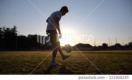 Professional footballer juggling soccer ball on stadium at sunset. Young man kicking ball at green field. Sportsman practicing tricks at meadow with sunlight at background. Freestyle football concept Professional footballer juggling soccer ball on stadium at sunset. Young man kicking ball at green field. Sportsman practicing tricks at meadow with sunlight at background. Freestyle football concept 122008285