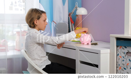 A young girl sits at her desk focused on placing coins into a piggy bank while writing notes about her savings. She learns the importance of managing personal finances A young girl sits at her desk focused on placing coins into a piggy bank while writing notes about her savings. She learns the importance of managing personal finances 122010203