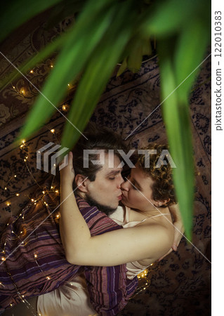 Couple kissing while lying on the carpet surrounded by flickering lights in a cozy room Couple kissing while lying on the carpet surrounded by flickering lights in a cozy room 122010383