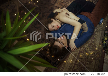 Couple embraces on a cozy rug surrounded by twinkling lights in a warm indoor setting Couple embraces on a cozy rug surrounded by twinkling lights in a warm indoor setting 122010384