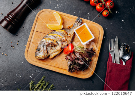 Grilled fish with lemon slices, salad, and dipping sauce on a wooden board for perfect seafood meal 122010647
