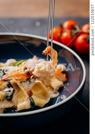 Close-up of a shrimp lifted with tongs from creamy pasta in a dark bowl 122010657