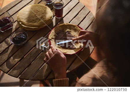 Person Spreading Chocolate Spread on Crepe at Wooden Table with Jam Nearby 122010730