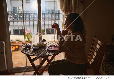Woman enjoying breakfast at a balcony table in warm sunlight Woman enjoying breakfast at a balcony table in warm sunlight 122010750