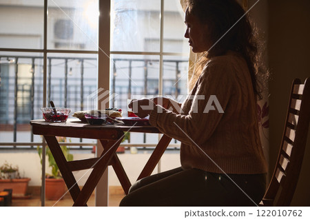 Woman enjoying a quiet meal with sunlight streaming through the window Woman enjoying a quiet meal with sunlight streaming through the window 122010762
