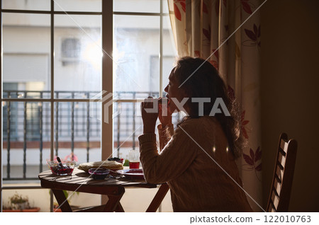 Woman enjoying breakfast near a sunlit window during a tranquil morning Woman enjoying breakfast near a sunlit window during a tranquil morning 122010763