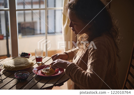 Woman enjoying crepes with jam and juice at home by window 122010764