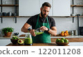 Man wearing a green apron pouring a green smoothie into a glass in a well-lit kitchen, surrounded by avocados and limes. 122011208