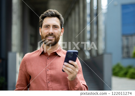 Portrait of successful businessman man with phone in hands. Office worker smiling and looking at camera, using app on smartphone, walking in city from outside office building. 122011274