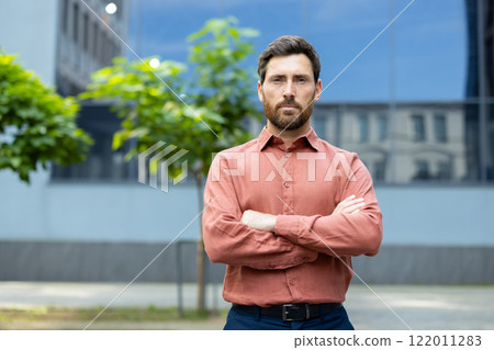 Portrait of serious successful office worker. Man looking at camera with folded hands outside office building. Businessman in red shirt confident and successful worker. 122011283
