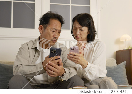 一對中年夫婦晚上在客廳一邊使用智慧型手機一邊擔心 122014263