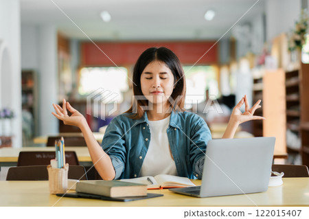 A young woman meditates at a desk in a library, surrounded by books and a laptop, embodying mindfulness and relaxation in a modern educational setting. 122015407