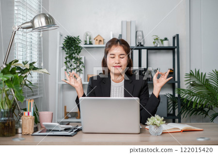 A businesswoman in a suit meditates at her desk in a modern office, surrounded by plants and a laptop, promoting mindfulness and stress relief. 122015420