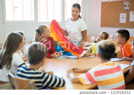 Children sitting in classroom and listening to female teacher. She holding rainbow flag in hands and talking about minorities Children sitting in classroom and listening to female teacher. She holding rainbow flag in hands and talking about minorities 122016332
