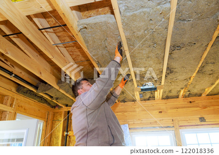 Worker is fitting insulation material into wooden ceiling beams of house, enhancing energy efficiency. Worker is fitting insulation material into wooden ceiling beams of house, enhancing energy efficiency. 122018336