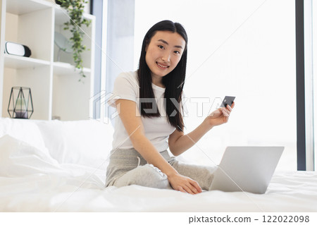 Asian woman sits on bed uses laptop for online shopping holding credit card. Casual home setting. Mid 20s enjoying convenience of internet shopping. 122022098