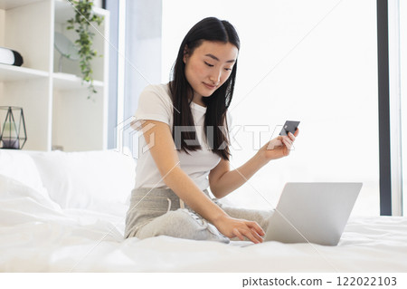 Young Asian woman makes online payment using laptop and credit card while sitting on bed. Relaxed and modern lifestyle setting. Young Asian woman makes online payment using laptop and credit card while sitting on bed. Relaxed and modern lifestyle setting. 122022103