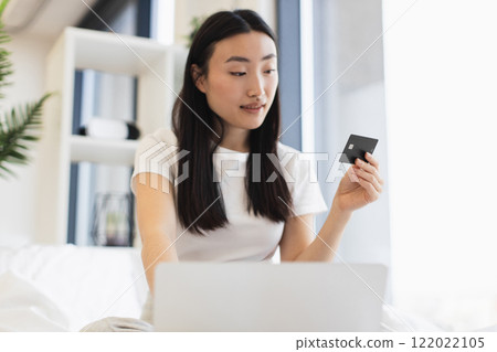 Young Asian woman makes online payment using laptop and credit card while sitting on bed. Relaxed and modern lifestyle setting. 122022105