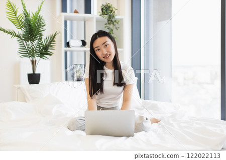 Young Asian woman sitting on bed using laptop in bright modern bedroom. She is smiling, creating relaxed and focused atmosphere, representing education and technology. Young Asian woman sitting on bed using laptop in bright modern bedroom. She is smiling, creating relaxed and focused atmosphere, representing education and technology. 122022113