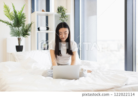Asian female in early twenties works on laptop in bright bedroom setting. Casual and relaxed atmosphere emphasizes work-life balance. 122022114