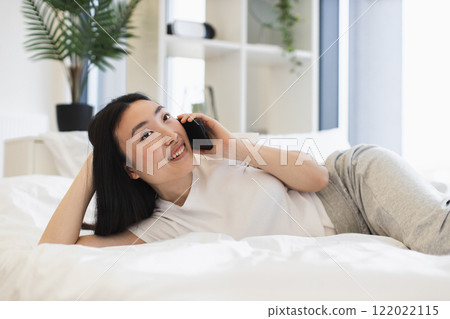 Young Asian woman smiling while having phone call on bed. She appears relaxed and content in a bright, modern bedroom setting. 122022115