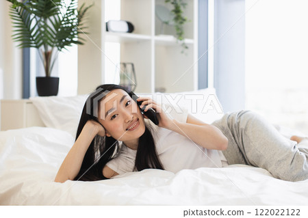 Smiling young Asian woman in casual attire lying on bed while talking on phone. Relaxed indoor setting. Comfortable and cheerful. Smiling young Asian woman in casual attire lying on bed while talking on phone. Relaxed indoor setting. Comfortable and cheerful. 122022122
