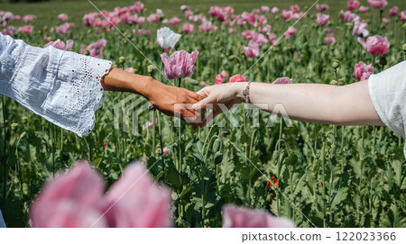 Two women's hands meet in a gentle touch, fingertips lightly brushing against each other amidst a field of pink poppies swaying gracefully in the wind.  122023366