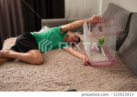 Young man resting on couch, holding a birdcage with a small green bird inside. 122023813
