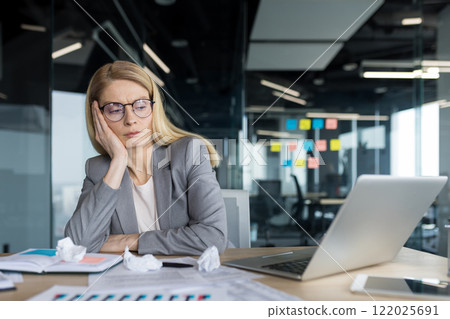 Tired and disappointed businesswoman sitting near documents and scraps of paper. Unsatisfied with results, woman in business suit received negative financial report results. 122025691