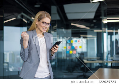 Mature businesswoman with phone in hands celebrating victory and successful achievement results. Woman at workplace read and received notification of victory and triumph online. Mature businesswoman with phone in hands celebrating victory and successful achievement results. Woman at workplace read and received notification of victory and triumph online. 122025723