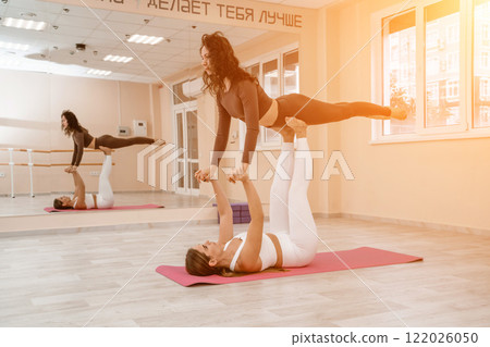 Two women do acroyoga on a light floor near a large mirror in a yoga studio. They are wearing leggings and tops. Two women do acroyoga on a light floor near a large mirror in a yoga studio. They are wearing leggings and tops. 122026050