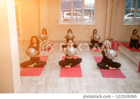 A group of six athletic women doing pilates or yoga on pink mats in front of a window in a beige loft studio interior. Teamwork, good mood and healthy lifestyle concept. 122026060