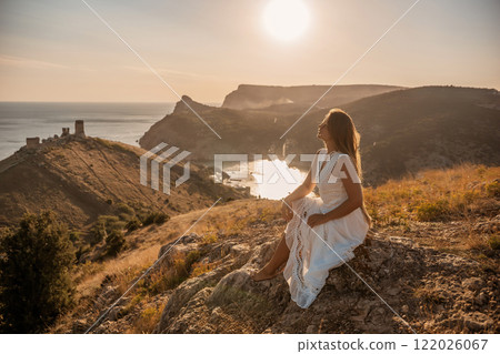 A woman in a white dress sits on a rock overlooking a body of water. The scene is serene and peaceful, with the woman enjoying the view and the calmness of the surroundings. 122026067