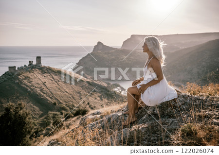 A woman is sitting on a hillside overlooking the ocean. She is wearing a white dress and has blonde hair. The scene is serene and peaceful, with the ocean in the background. A woman is sitting on a hillside overlooking the ocean. She is wearing a white dress and has blonde hair. The scene is serene and peaceful, with the ocean in the background. 122026074