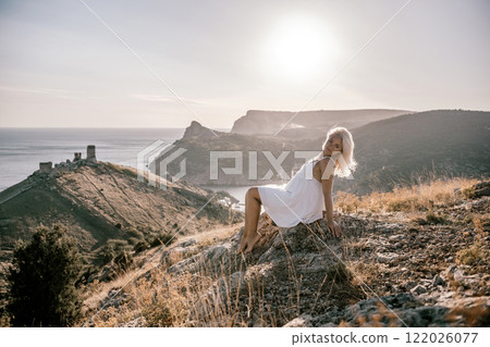 A woman is sitting on a hillside overlooking the ocean. She is wearing a white dress and has blonde hair. The scene is serene and peaceful, with the ocean in the background. A woman is sitting on a hillside overlooking the ocean. She is wearing a white dress and has blonde hair. The scene is serene and peaceful, with the ocean in the background. 122026077