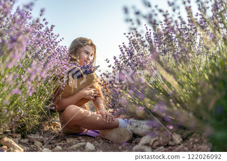 Girl is sitting in a field of purple flowers. She is holding a basket of flowers and smiling. Scene is peaceful and serene, as the girl is surrounded by the beauty of nature. Girl is sitting in a field of purple flowers. She is holding a basket of flowers and smiling. Scene is peaceful and serene, as the girl is surrounded by the beauty of nature. 122026092