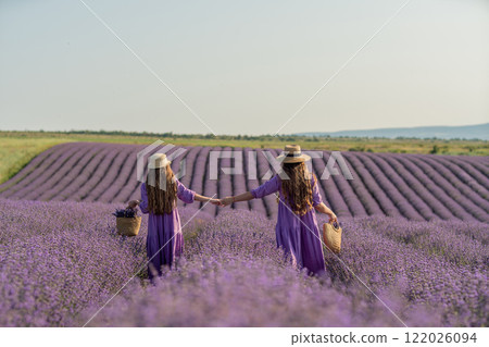 Mom and daughter are running through a lavender field dressed in purple dresses, long hair flowing and wearing hats. The field is full of purple flowers and the sky is clear. Mom and daughter are running through a lavender field dressed in purple dresses, long hair flowing and wearing hats. The field is full of purple flowers and the sky is clear. 122026094