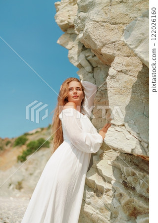 A woman in a white dress is standing on a rocky beach. Concept of serenity and tranquility, as the woman is enjoying the natural beauty of the beach. 122026160