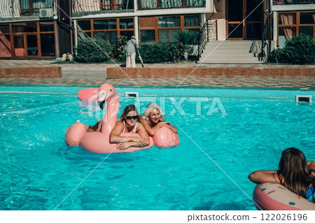 Three women are floating in the pool on inflatable floats. One of the floats is pink and has a flamingo on it. The women are smiling and enjoying their time in the water. 122026196