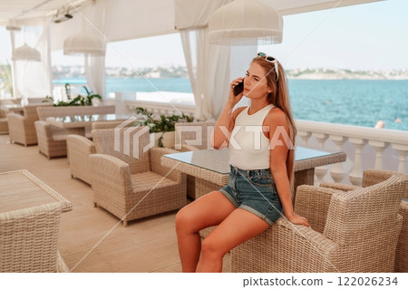 Cafe woman sitting at a table with a cell phone in her hand. She is looking at the screen and she is focused on her phone. The scene takes place in a restaurant or cafe, with multiple chairs. 122026234
