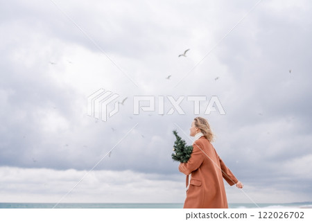 Blond woman Christmas tree sea. Christmas portrait of a happy woman walking along the beach and holding a Christmas tree on her shoulder. She is wearing a brown coat and a white suit. 122026702