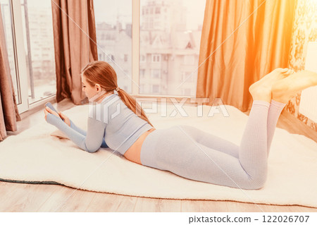 Side view portrait of relaxed woman with long hair lying on carpet at home. She is dressed in a blue tracksuit, holding a phone in her hands. Side view portrait of relaxed woman with long hair lying on carpet at home. She is dressed in a blue tracksuit, holding a phone in her hands. 122026707