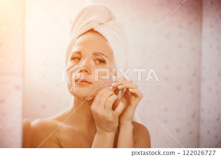 Young beautiful woman using a moisturizing facial mask after taking a bath. Pretty attractive girl in a towel on her head stands in front of a mirror in a home bathroom. Daily hygiene and skin care Young beautiful woman using a moisturizing facial mask after taking a bath. Pretty attractive girl in a towel on her head stands in front of a mirror in a home bathroom. Daily hygiene and skin care 122026727