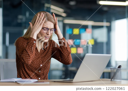 Businesswoman anxiously holding hands on head, received negative achievement results, bankrupt. Desperate woman working at desk inside office, using notebook. Businesswoman anxiously holding hands on head, received negative achievement results, bankrupt. Desperate woman working at desk inside office, using notebook. 122027061