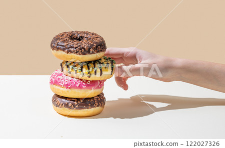 Hand takes one donut from stack of glazed donuts with different toppings on beige background. 122027326