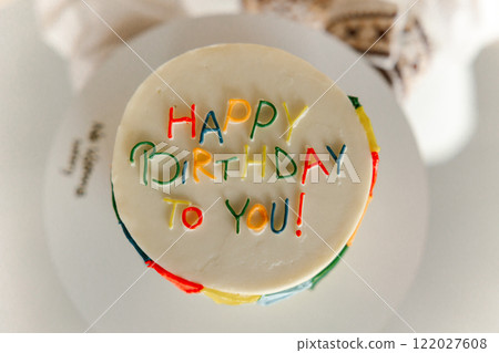 Top view of festive round white cream cake with bright colourful "Happy Birthday to you" inscription on stand on table. Birthday celebration, dessert, cooking, greetings. Party concept. Top view of festive round white cream cake with bright colourful "Happy Birthday to you" inscription on stand on table. Birthday celebration, dessert, cooking, greetings. Party concept. 122027608