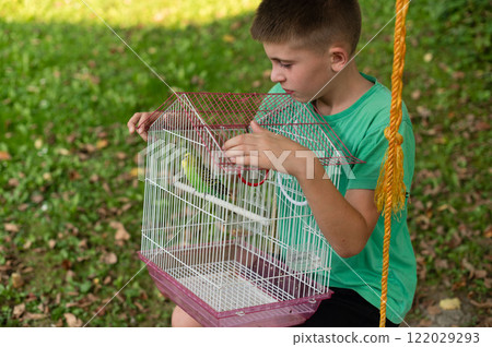 Young boy gently holds a birdcage containing a small green bird outdoors. 122029293