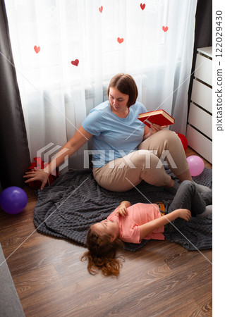 Caucasian young mother reads a book to her daughter, sitting on the floor by the window. Preschool education at home. Vertical 122029430