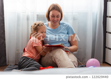 Mid shot of young Caucasian woman reads a book of fairy tales to her child. A preschool-age cute girl is interested in spending time with her mother. Concept of mother's day Mid shot of young Caucasian woman reads a book of fairy tales to her child. A preschool-age cute girl is interested in spending time with her mother. Concept of mother's day 122029440
